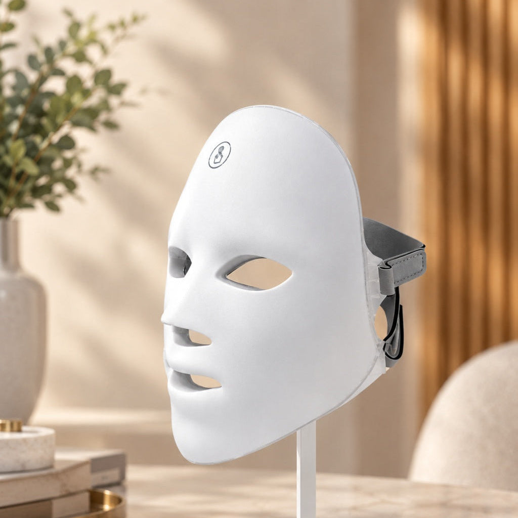 A white LED facial mask therapy device on a white stand, set on a marble table in a warm, modern room with a plant and curtains in the background.