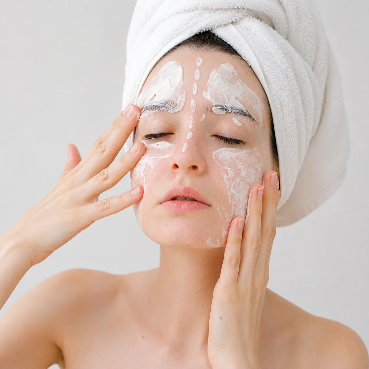 A woman with a white towel wrapped around her hair applies a white facial cleanser or mask cream to her face with her hands, set against a plain light gray background.
