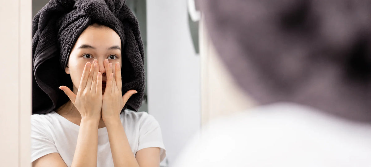 A woman with a dark towel wrapped around her hair looks into a mirror while applying or washing her face with her hands. She is wearing a white t-shirt in a bathroom setting.